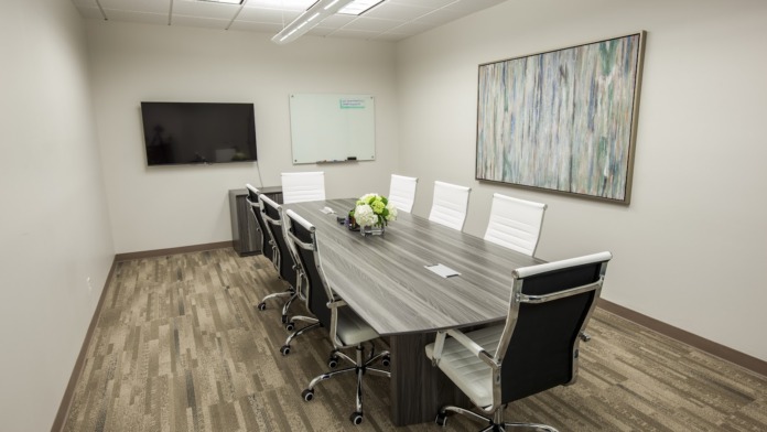 Interior meeting space with long gray conference table, white office chairs, mounted TV, and whiteboard