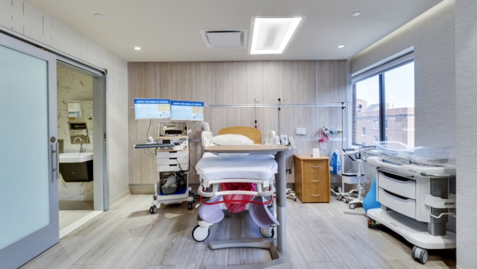 Interior patient treatment room with hospital bed, medical equipment, wooden cabinetry, and contemporary flooring.
