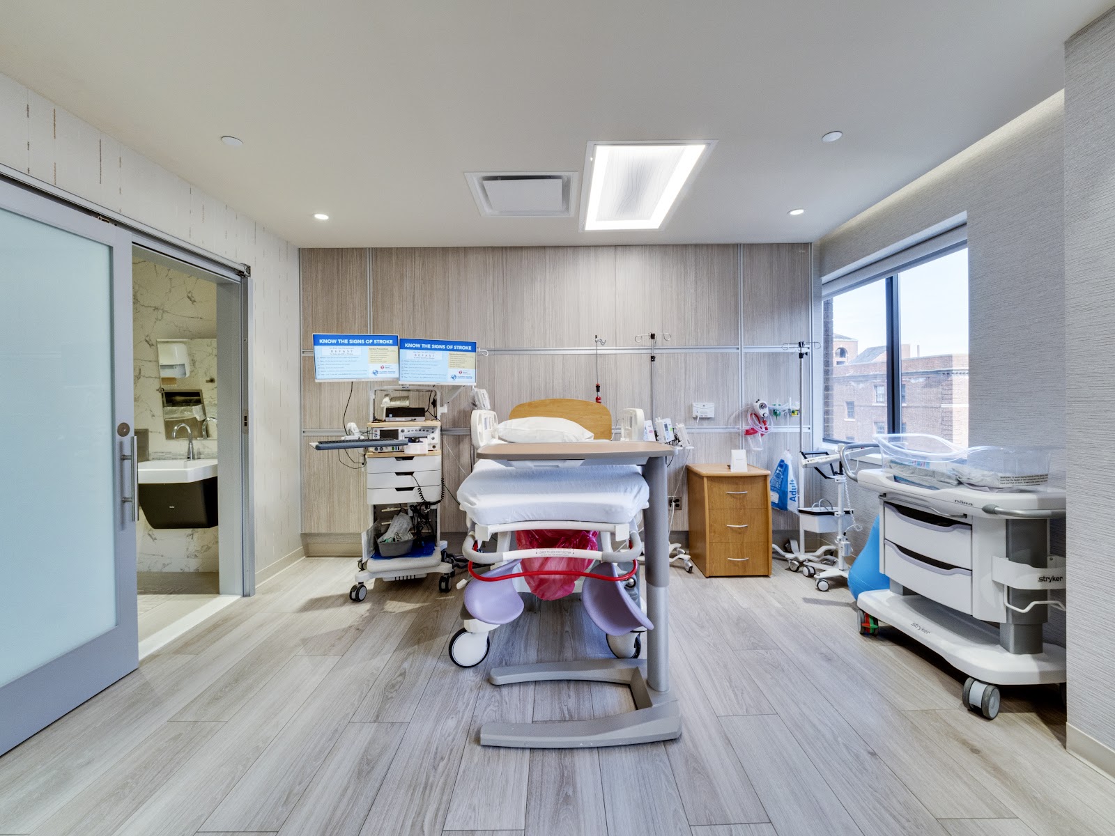 Interior patient treatment room with hospital bed, medical equipment, wooden cabinetry, and contemporary flooring.