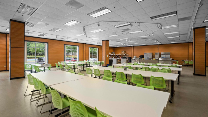 Interior room with warm orange wood paneling, white ceiling, lime green chairs, long white tables, and large windows