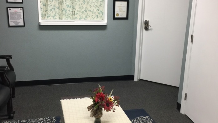 Interior meeting room with light blue walls, framed documents, window with patterned curtains, and wooden table with flower arrangement