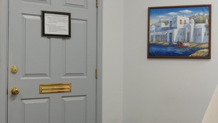Interior office space with gray six-panel door, information plaque, framed coastal painting, wooden bench, and warm lighting