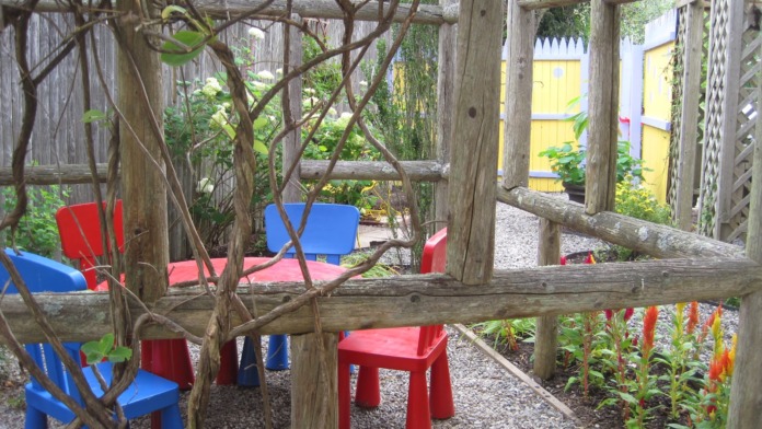Outdoor pergola with wooden beams, climbing vines, red and blue children's furniture, and flowering plants