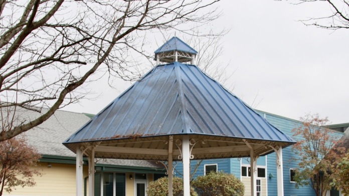 Exterior outdoor gazebo with blue metal roof, white support columns, and multiple lounge chairs on brick patio surrounded by landscaping