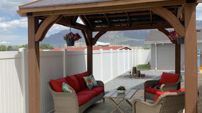 Covered outdoor patio with wooden pergola structure, red cushioned seating, wicker chairs, stone pathway, and mountain views.