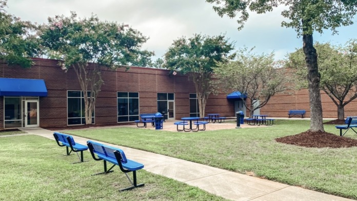 Outdoor patio with blue picnic tables, brick building, mature landscaping, lawn, and trees creating gathering space