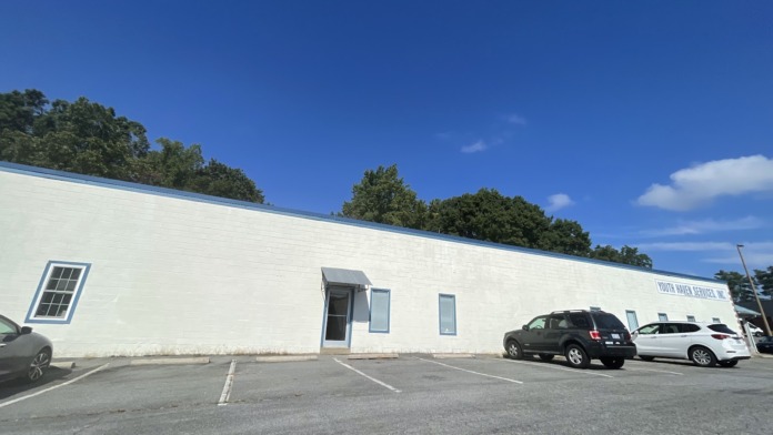 Exterior view of white industrial building with clear blue sky, parking area with vehicles, and tree line background