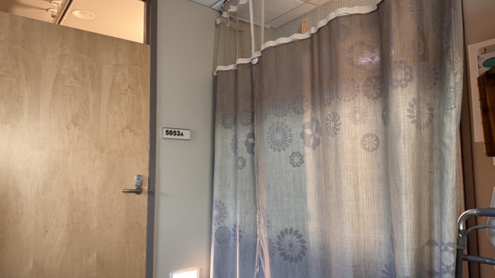 Interior patient bathroom showing toilet, shower with patterned curtain, tile flooring, and room identification.