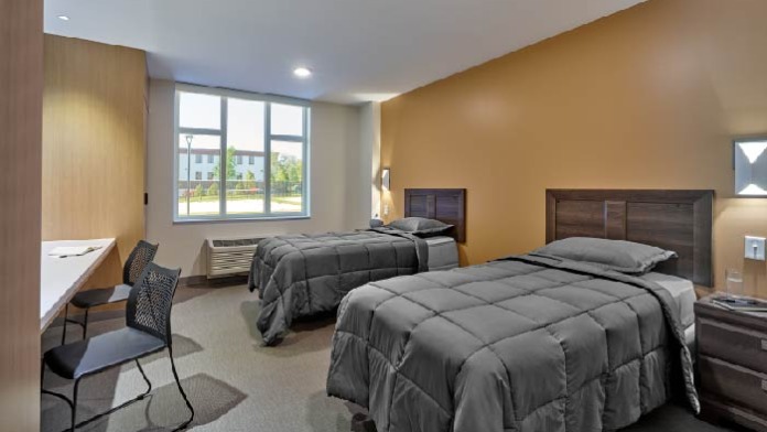 Interior patient bedroom with two queen beds, desk area, wooden furniture, and windows with natural lighting.