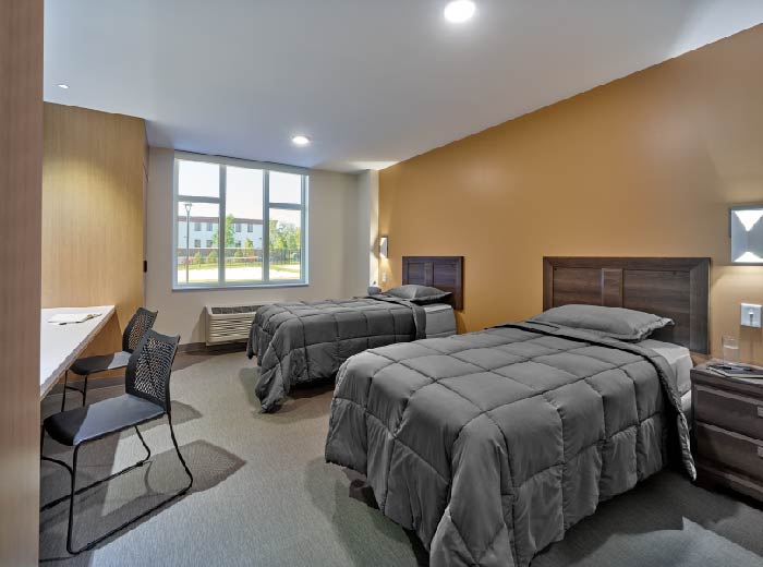 Interior patient bedroom with two queen beds, desk area, wooden furniture, and windows with natural lighting.