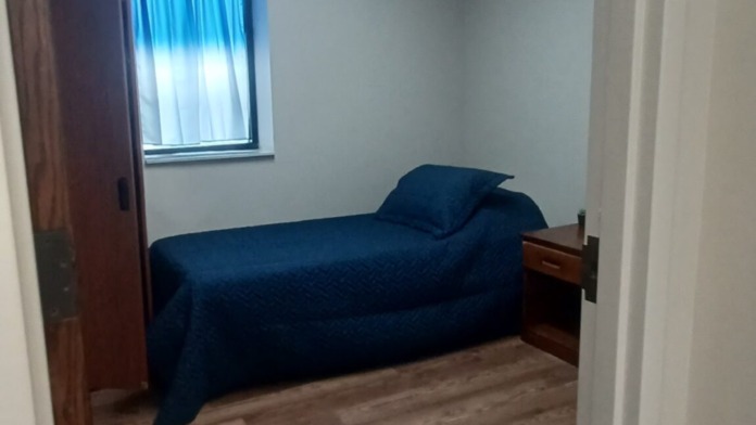 Interior bedroom viewed through doorway showing single bed with dark bedding, nightstand, window, and wooden flooring.