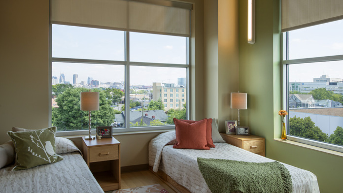 Interior patient bedroom with twin beds, green accent walls, city view windows, and wooden furniture.