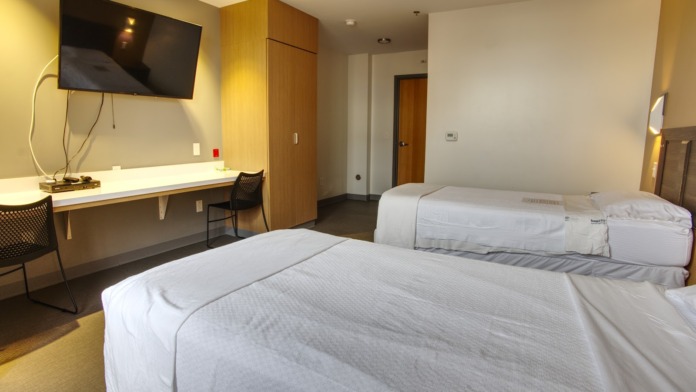 Interior patient bedroom with two beds, desk with monitor, wooden storage cabinetry, and warm overhead lighting.