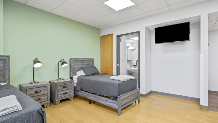 Interior patient bedroom featuring two beds with gray bedding, wooden nightstands, green accent wall, and en-suite bathroom