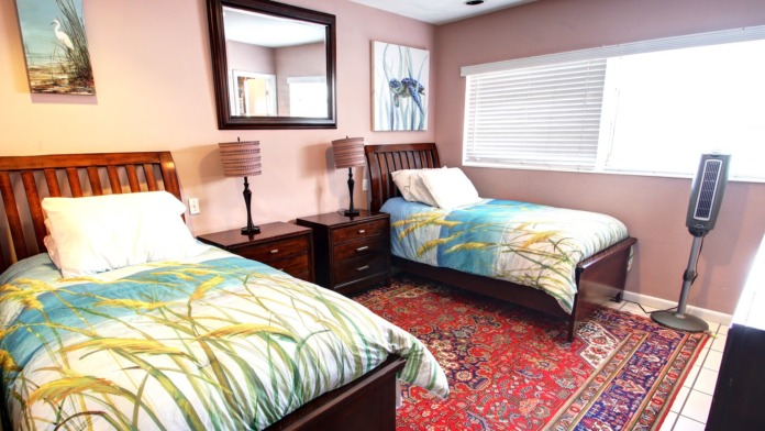 Interior patient bedroom showing two twin beds with matching bedding, pink walls, nightstands, large window, and area rug