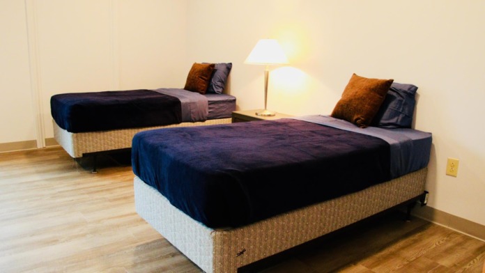 Interior patient bedroom featuring two twin beds with dark blue bedding, decorative pillows, bedside lamp, and wooden flooring