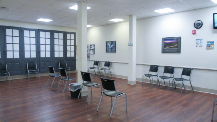 Interior waiting room with rows of metal-frame chairs, dark glass doors, wood flooring, and framed wall decorations