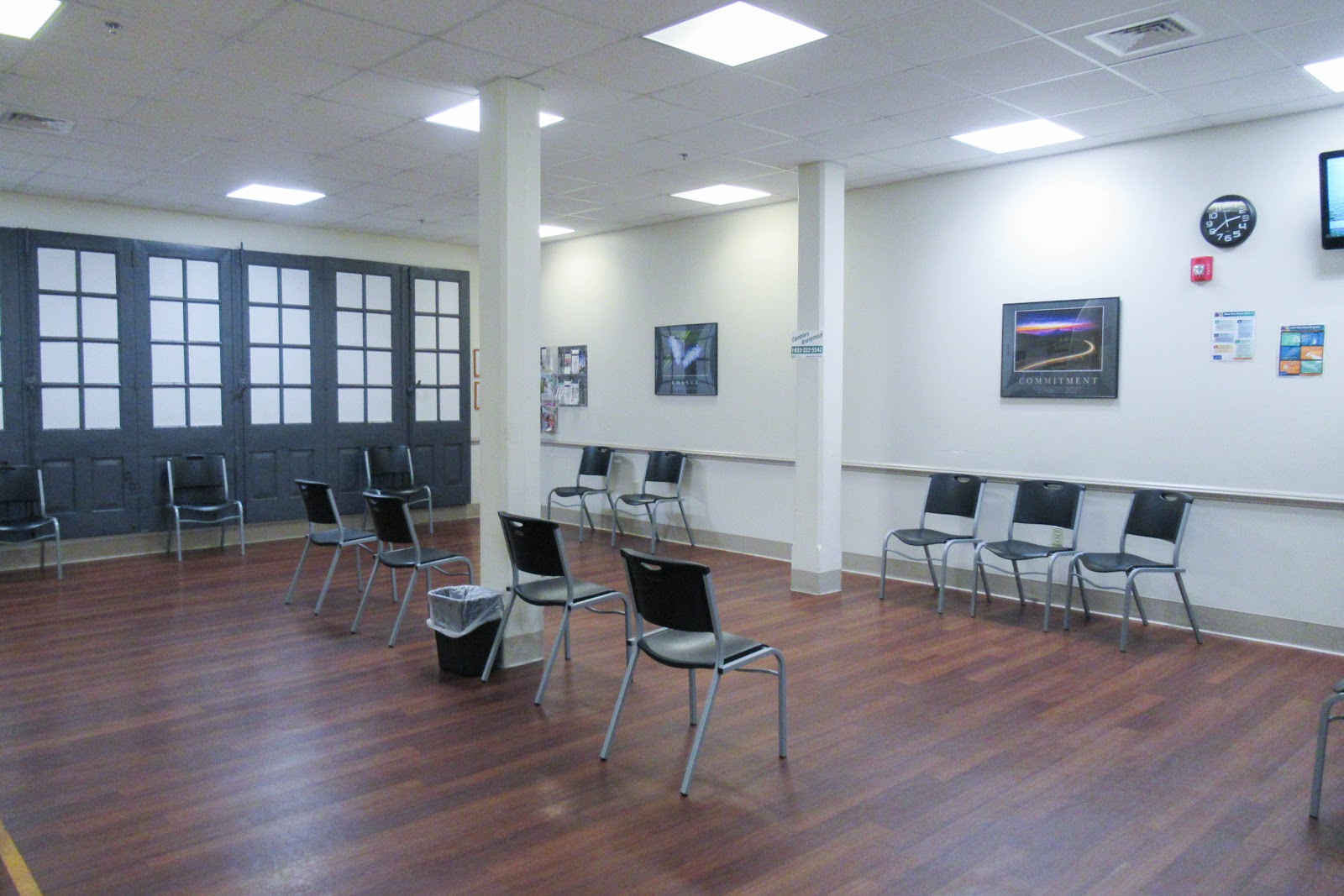 Interior waiting room with rows of metal-frame chairs, dark glass doors, wood flooring, and framed wall decorations