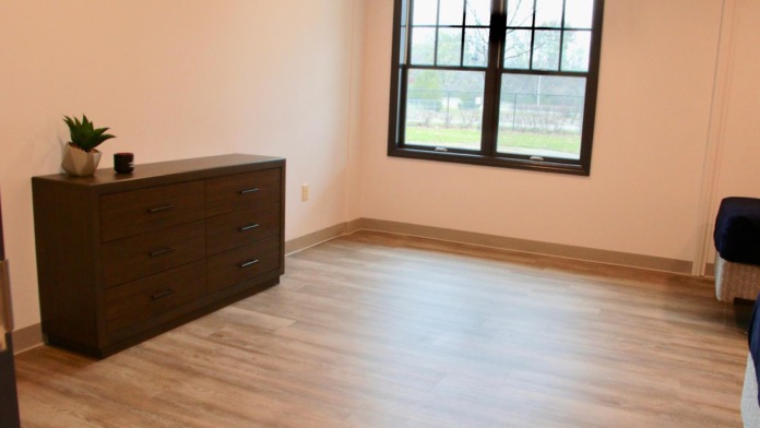 Interior bedroom showing wooden dresser, large window with black frame overlooking grounds, hardwood flooring, and minimal furnishings