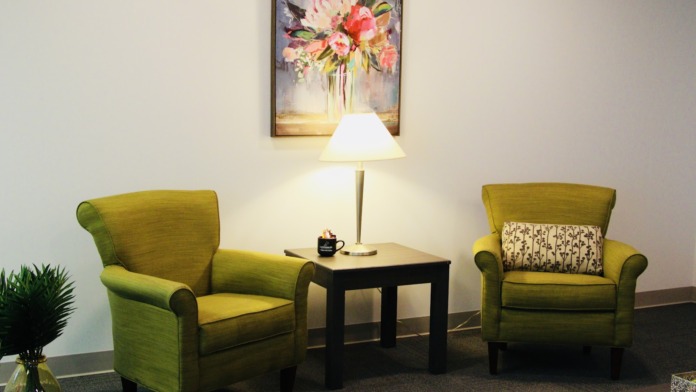 Interior seating area with two green upholstered chairs, black side table, table lamp, framed floral artwork on wall, and potted plant