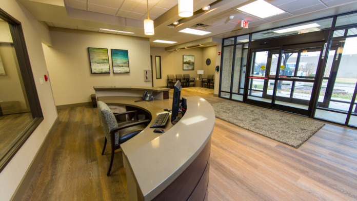 Interior reception lobby with curved white counter desk, chairs, landscape artwork, glass entry doors, wood flooring.