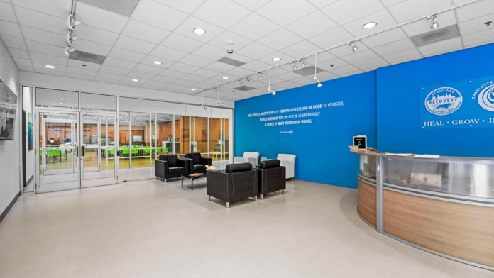 Interior lobby with blue accent wall, facility logo signage, black seating, wooden reception desk, and glass entry doors
