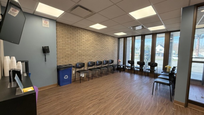Interior reception space with tan and brick walls, wooden flooring, seating chairs, counter, and large windows.