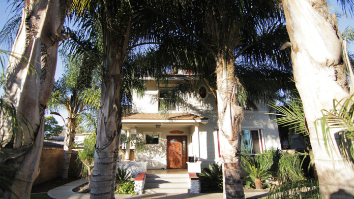 Two-story white building with multiple tall palm trees, brick accents, fencing, and neighboring residential properties.