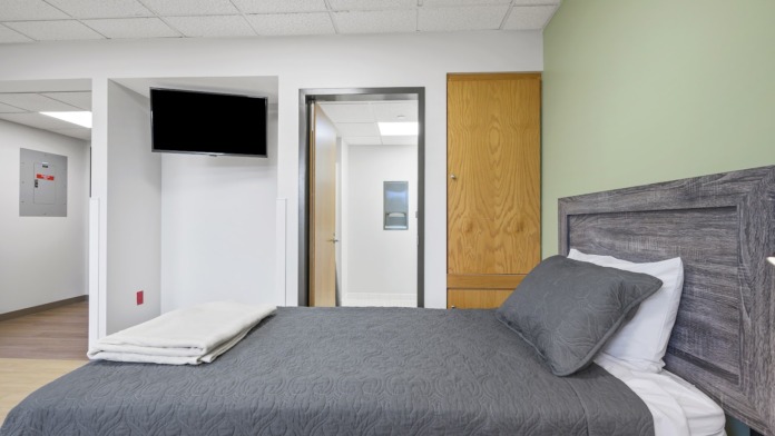 Interior patient bedroom with queen bed, gray bedding, wooden headboard, mounted TV, and wooden flooring