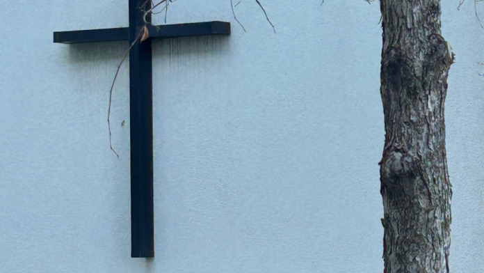 Exterior wall featuring mounted black cross with stone foundation and bare tree branches overhead.