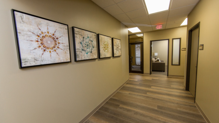 Interior hallway with yellow walls, framed abstract artwork, office doors, wooden flooring, and overhead lighting.
