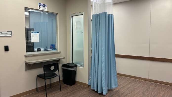 Interior treatment area with examination room behind glass partition, curtained privacy screen, and office window