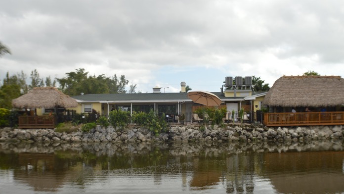 Exterior view of facility from waterfront showing yellow and tan buildings with thatched roof structures, wooden decking, and water reflection