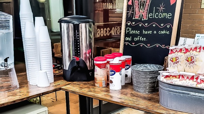 Interior refreshment station with coffee maker, welcome sign, cups, and snacks on wooden table
