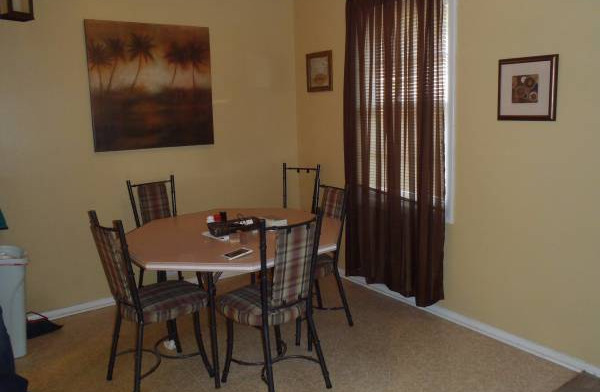 Interior dining room with round table, yellow and blue walls, kitchen visible, and natural window light