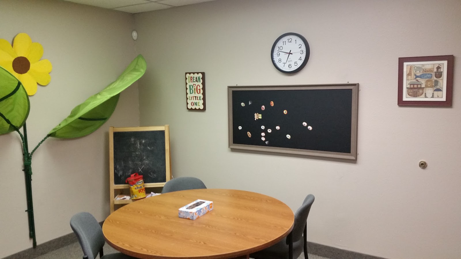 Interior space with wooden round table, decorative floor flowers, wall clock, and colorful framed artwork.