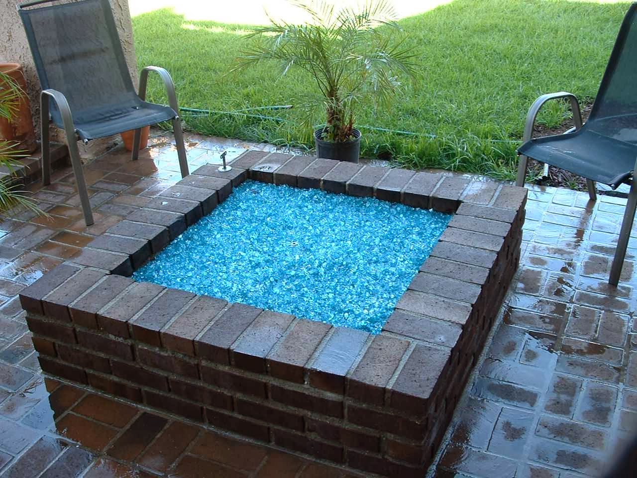 Backyard fire pit with blue glass rocks, brick seating surround, metal chairs, and landscaped lawn area