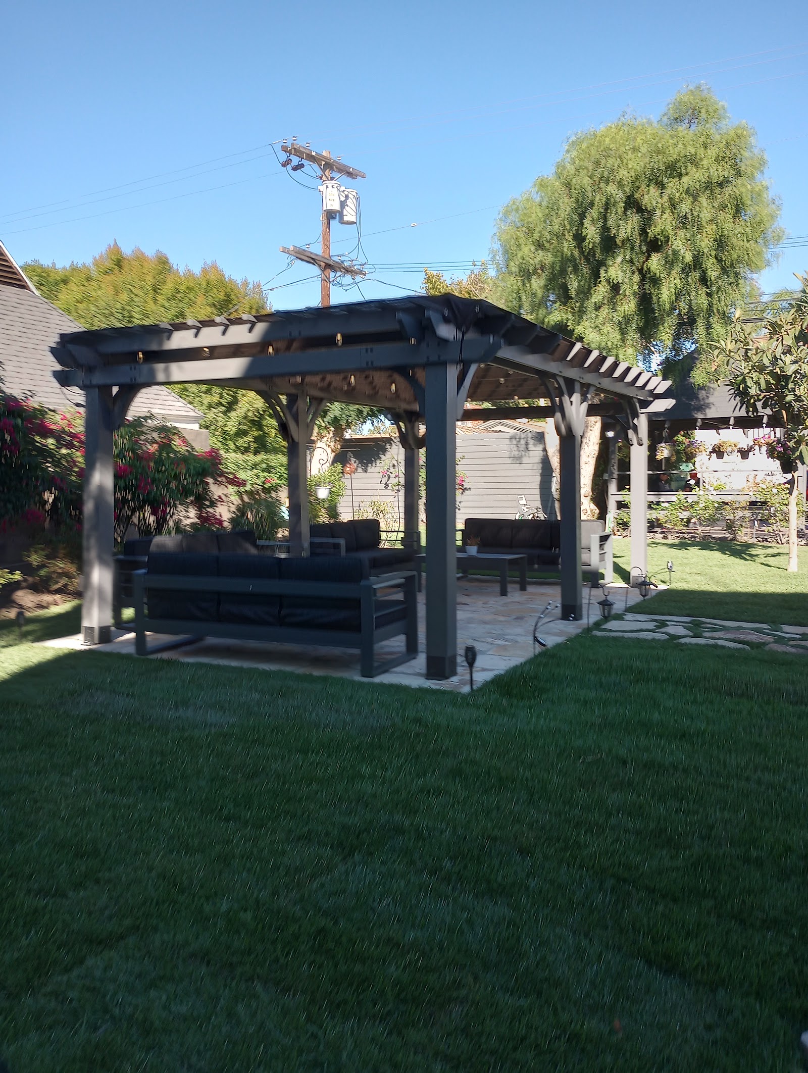 Outdoor seating area with pergola structure, comfortable seating, manicured lawn, flowering plants and trees