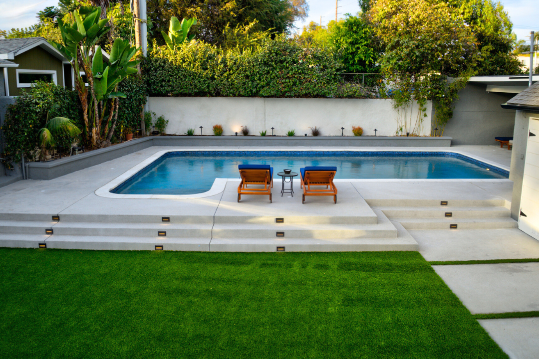 Outdoor swimming pool with modern concrete deck, lounge chairs, mature landscaping, and privacy walls.