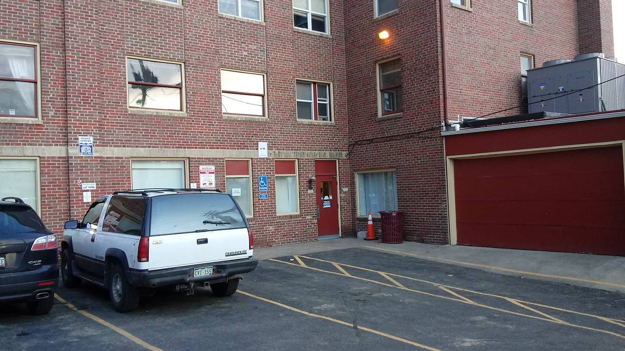 Exterior view of red brick facility building with multiple windows, parking area with vehicles, and entrance