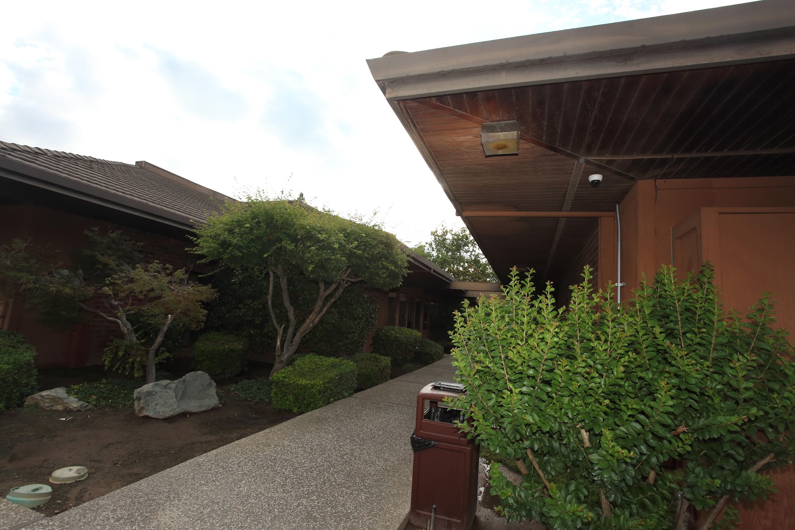 Exterior view of brick building with mature trees, landscaped grounds, gravel pathway, and covered structure with wooden ceiling