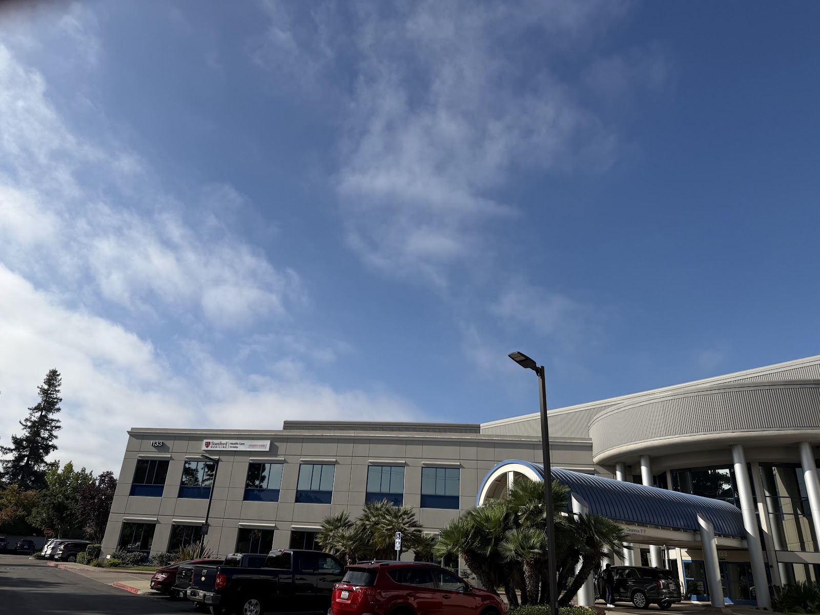 Exterior view of modern medical facility with gray and tan facade, parking lot with landscaping, and clear blue sky