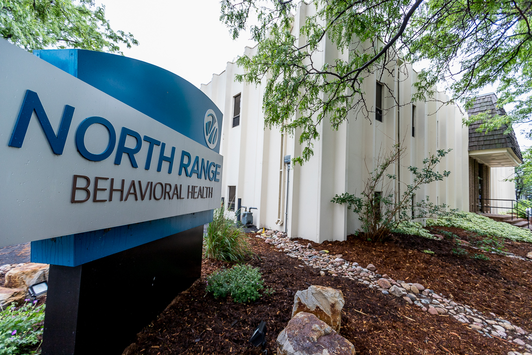 Exterior showing North Range Behavioral Health sign, building facade with windows, landscaping, and mulched grounds.
