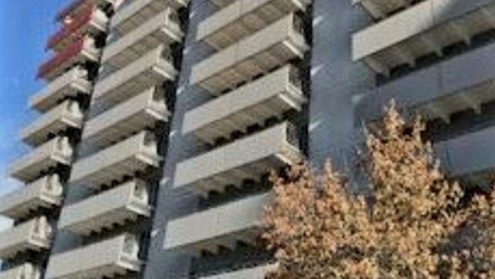 Exterior view of multi-story residential building with white facade, horizontal balconies, trees, and parking area visible