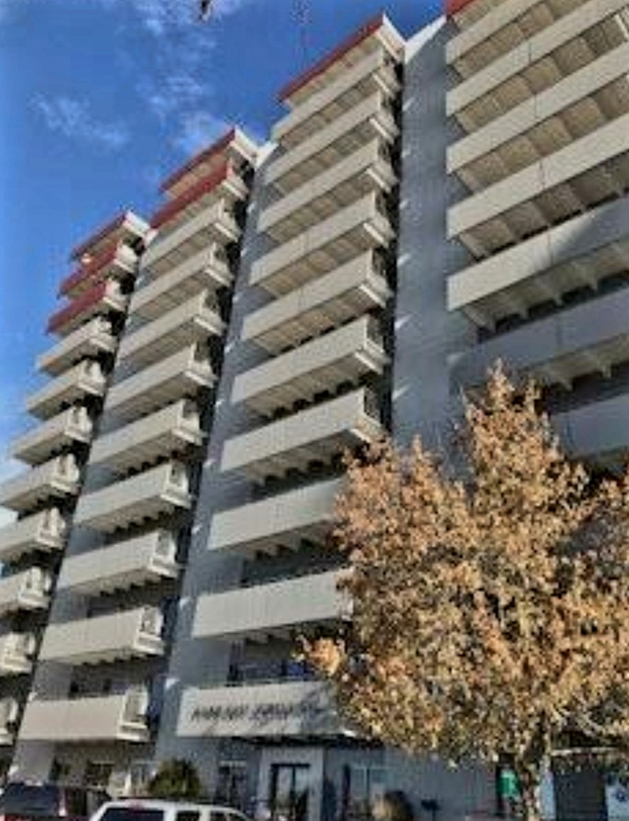 Exterior view of multi-story residential building with white facade, horizontal balconies, trees, and parking area visible