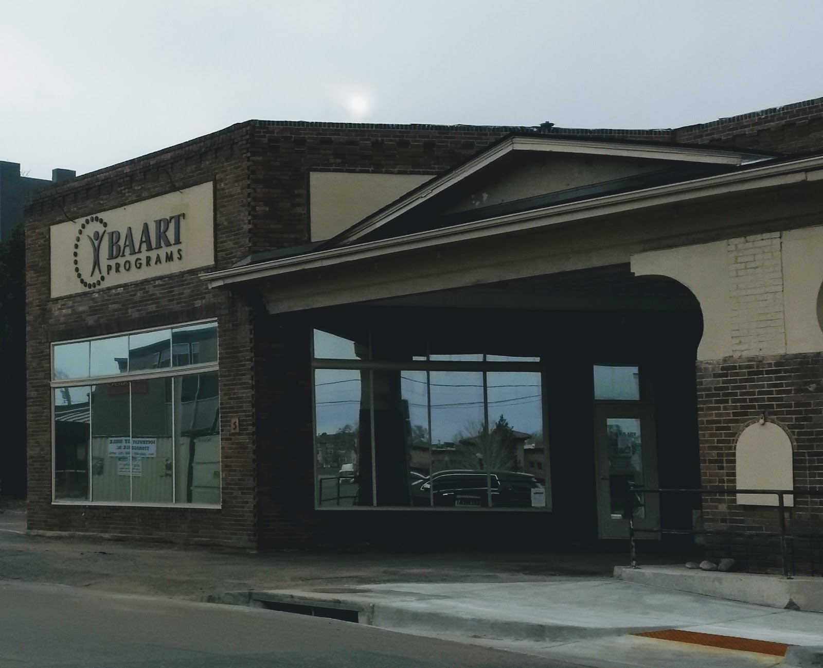 Exterior brick building facade with BAART Programs signage visible, large glass window, cloudy sky