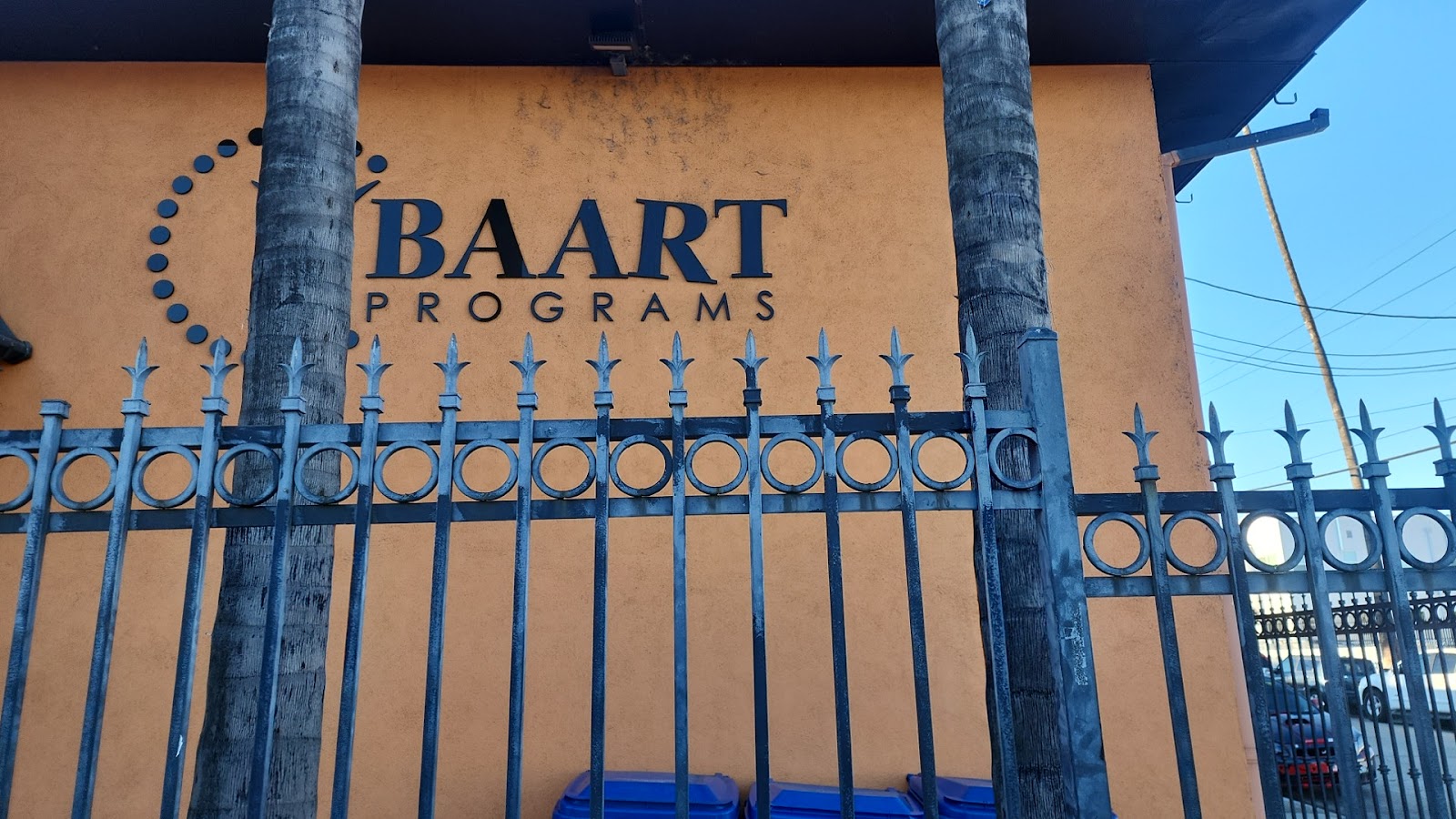 Exterior close-up of BAART Programs signage on orange building wall with decorative blue metal fence and palm tree trunks