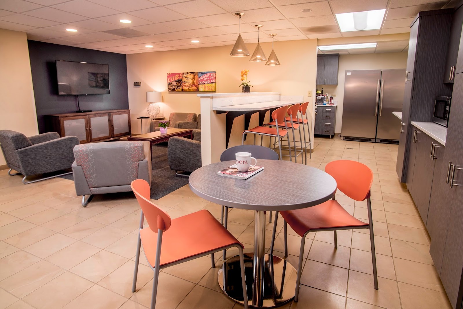 Interior community space with round tables, orange chairs, bar seating, kitchen area, and televised entertainment