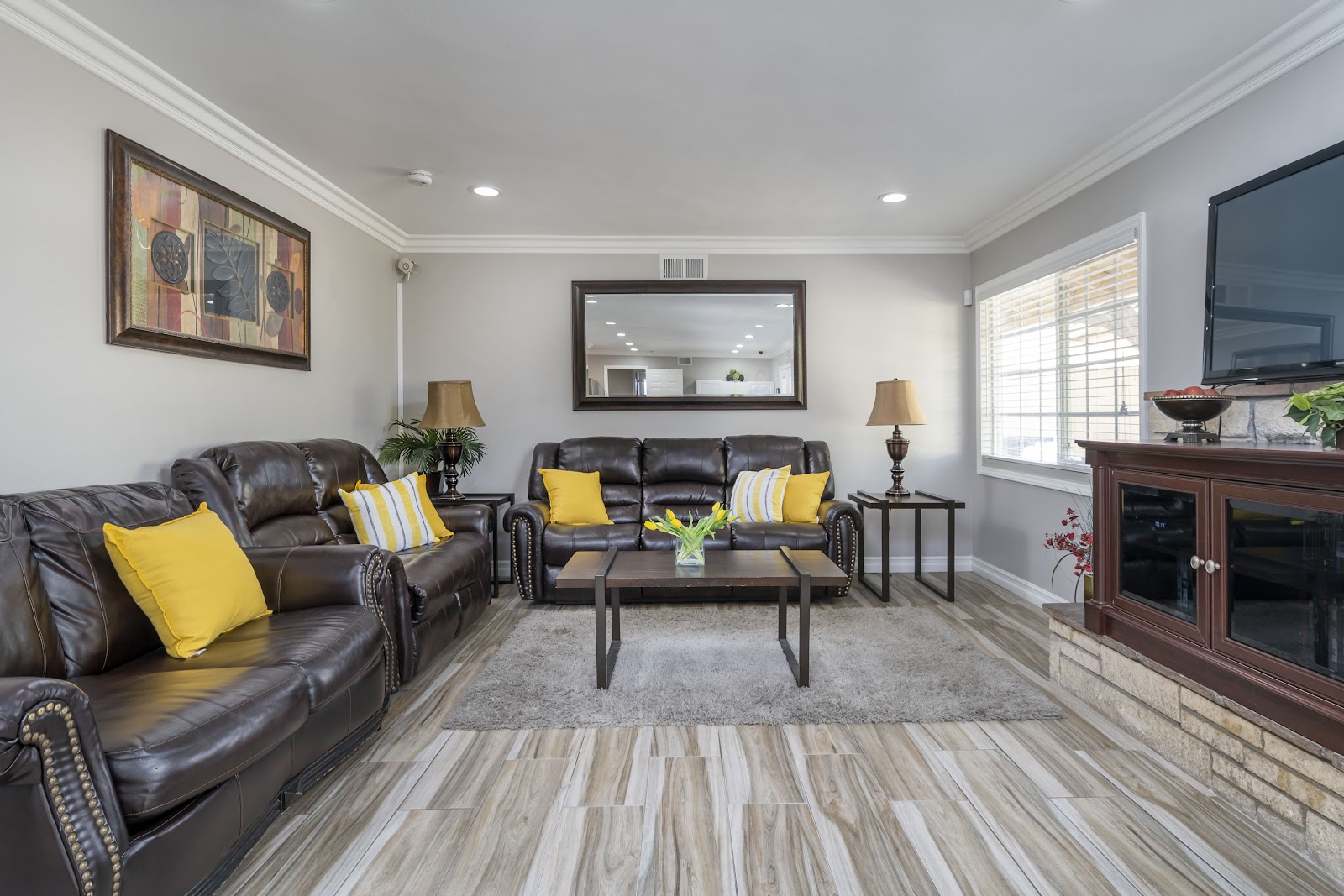 Interior common area with leather seating, yellow accent pillows, fireplace, framed artwork, mirror, television, and large windows.