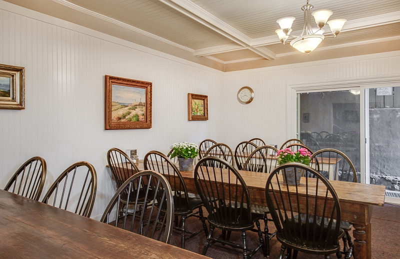 Interior dining area with long wooden table, black Windsor-style chairs, framed landscape artwork, windows with shutters, and pendant lighting